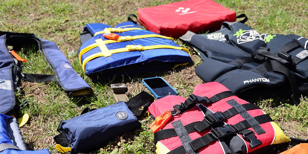Various types of life jackets laid out on grass: Throwable, powerboating life jacket, standard life jacket, youth life jacket and two inflatable life jackets