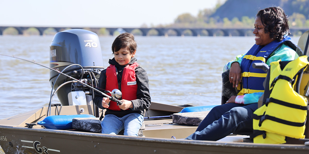 A young boy and a woman sit on a boat wearing life jackets on the Susquehanna River. The young boy is holding his fishing rod and reeling in his line.