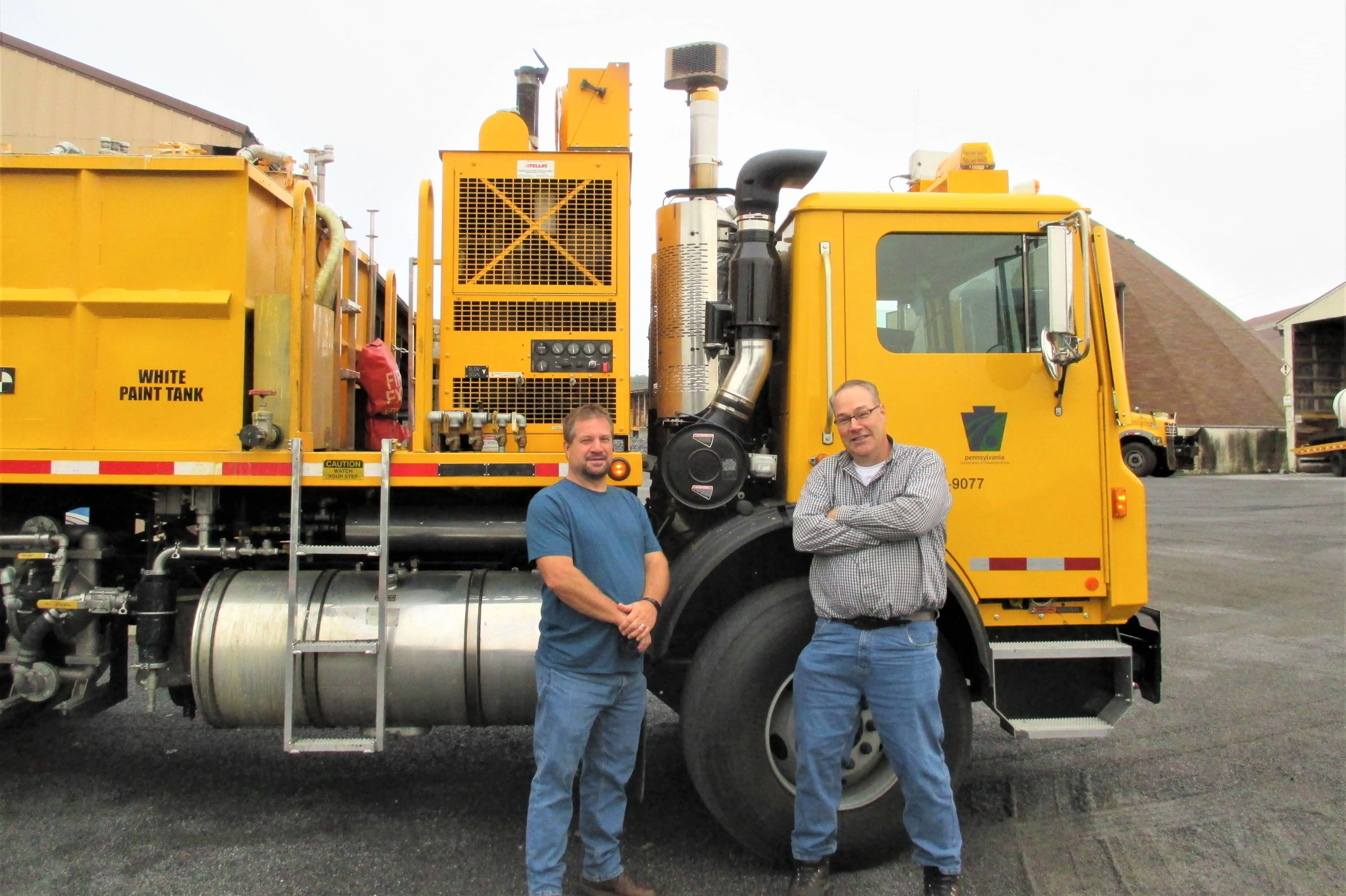 Two employees pose in front of the cab of a PennDOT paint truck carrying a paint tank filled with white line paint.