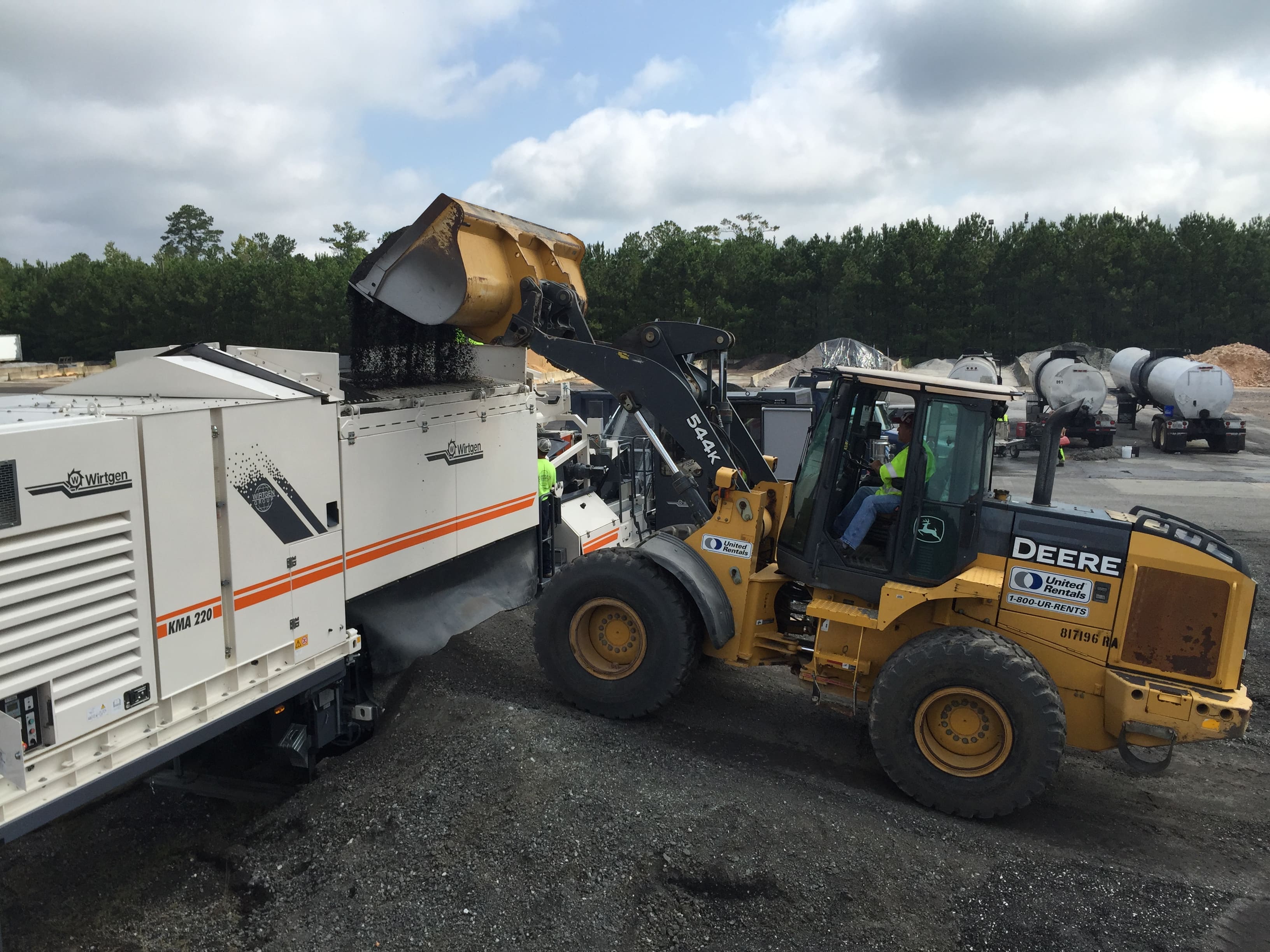 An image of a worker operating a backhoe to place Reclaimed Asphalt Pavement (RAP) in a white, rectangle-shaped, metal mobile cold recycling plant where it is mixed with a liquid, such as asphalt emulsion or foamed asphalt, and a small amount of cement to create a new flexible asphalt base.