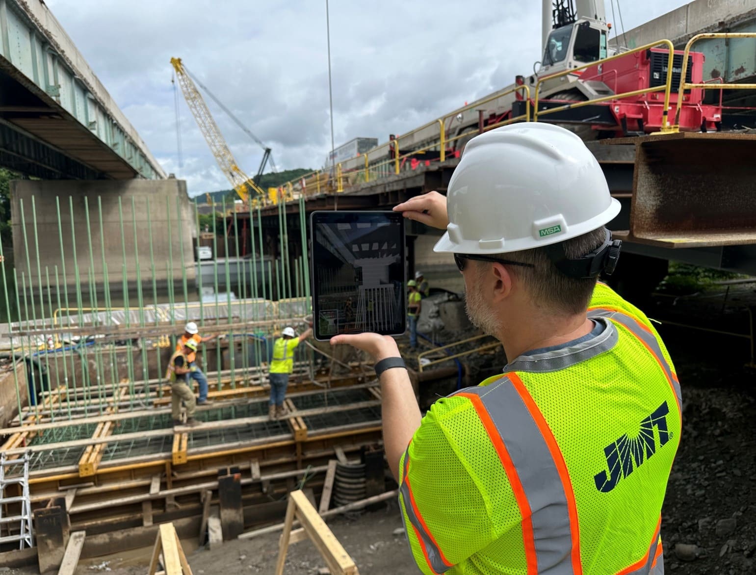 A man wearing a white hard hat, yellow safety vest and sunglasses stands at a bridge construction site holding tablet.