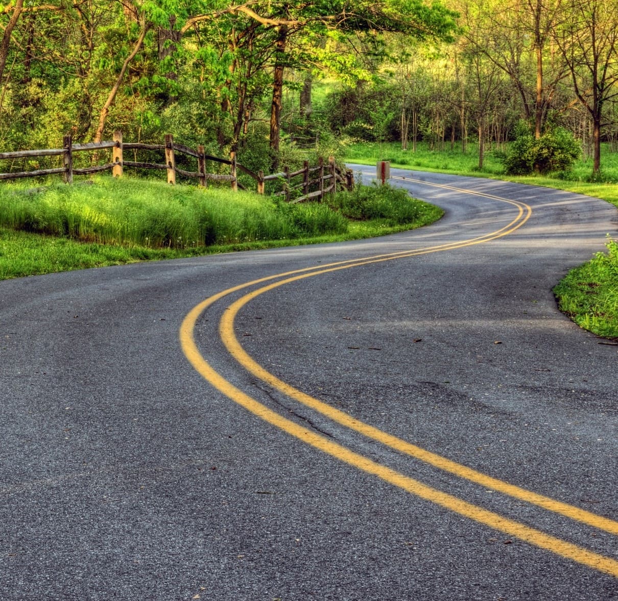An image of a two-lane, winding roadway, lined with grass, trees and a fence.