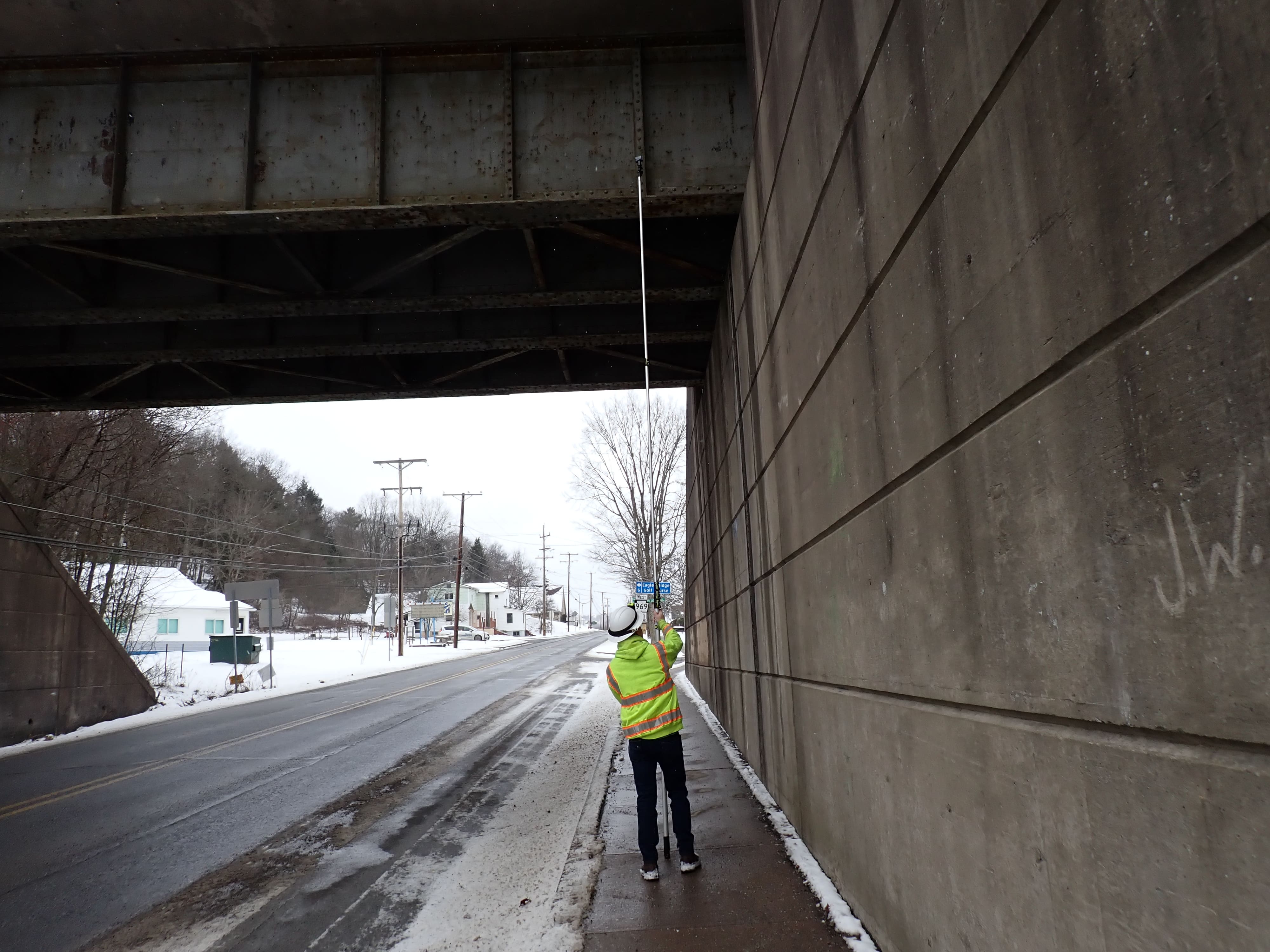 An individual in a yellow safety vest and white hard hat holds a pole with a GoPro attached underneath a bridge.
