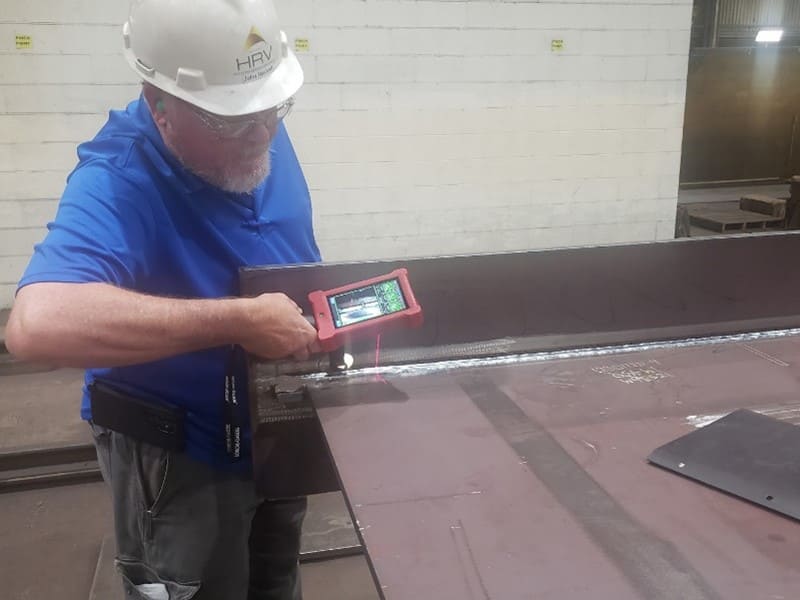 A man wearing a blue shirt, safety goggles and a white hard hat holds up a scanning device next to a piece of metal.