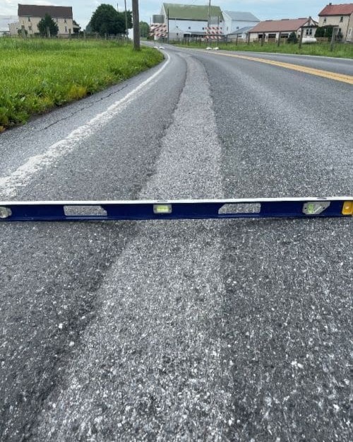 An image of a metal level sitting in the middle of a trough that has developed on the roadway. Grass, trees and houses are in the background of the photo.