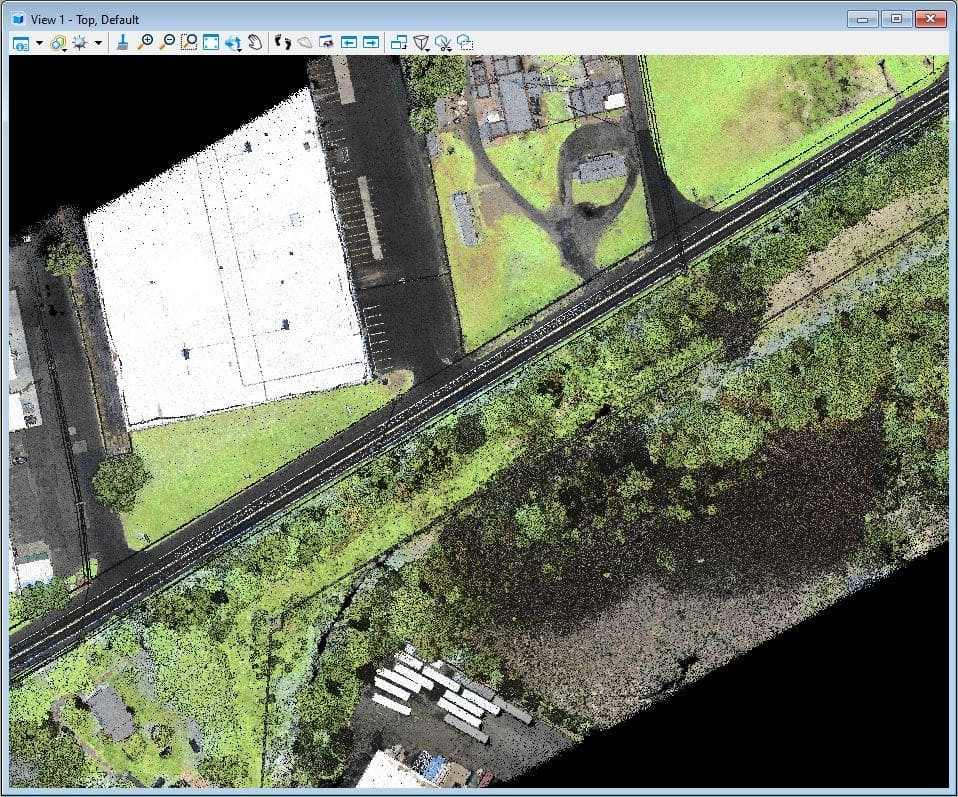 An image of a two-lane roadway, with a large warehouse on the left of the roadway and a large number of trees and other foliage to the right of the roadway.