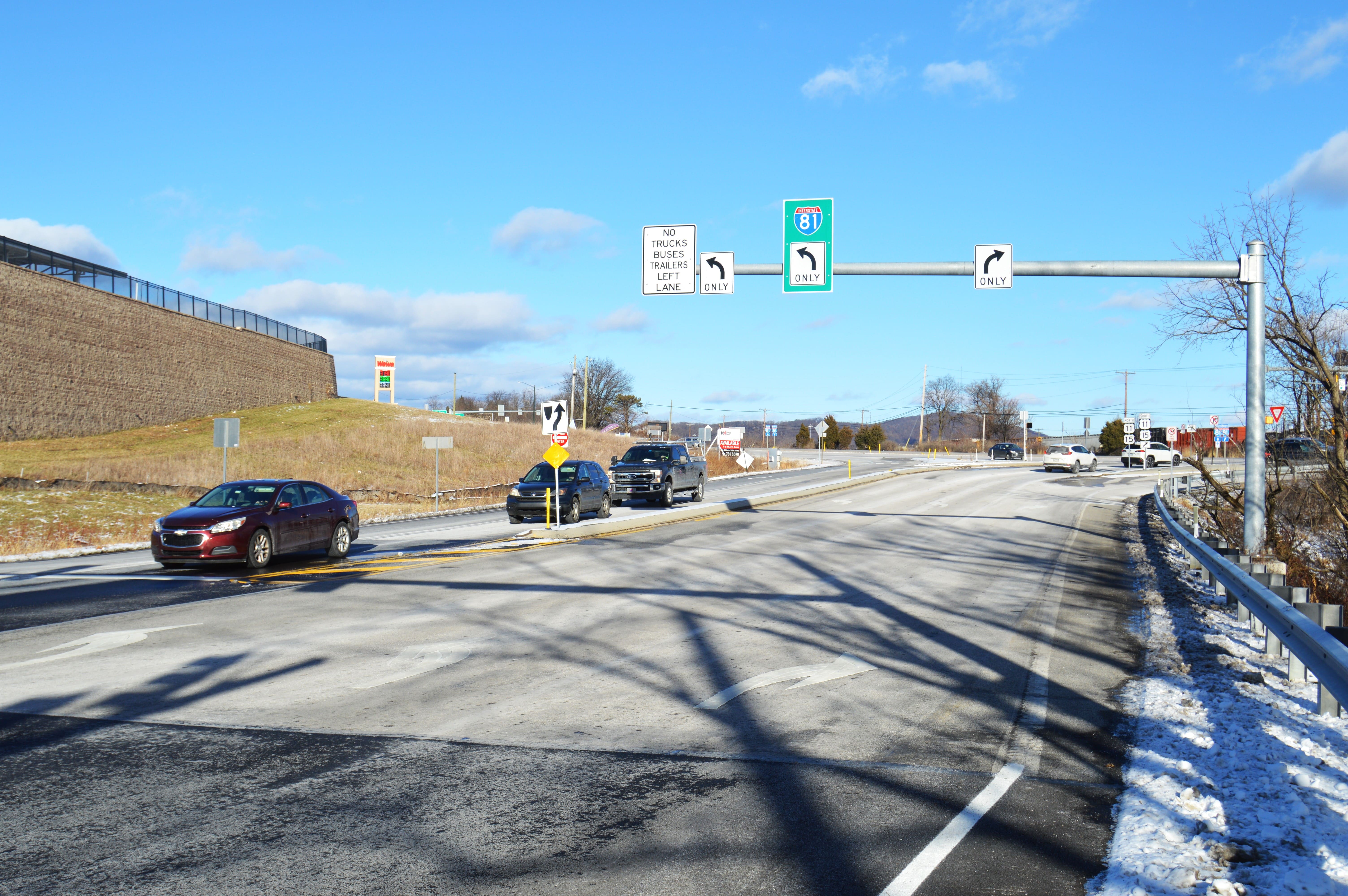A red car, black SUV and black pickup truck passing through an intersection. On the left, there are overhead directional signs for opposing traffic.  