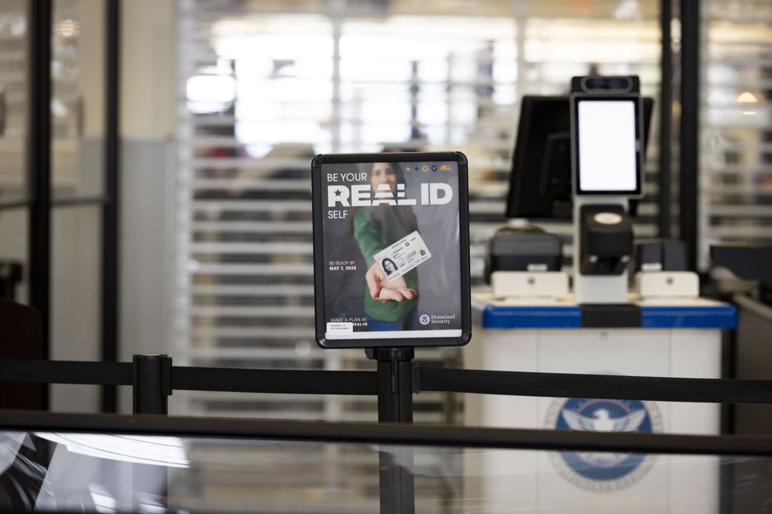 A Department of Homeland Security sign that says 'Be Your REAL ID Self' sits atop a black stanchion at a commercial airport security checkpoint.
