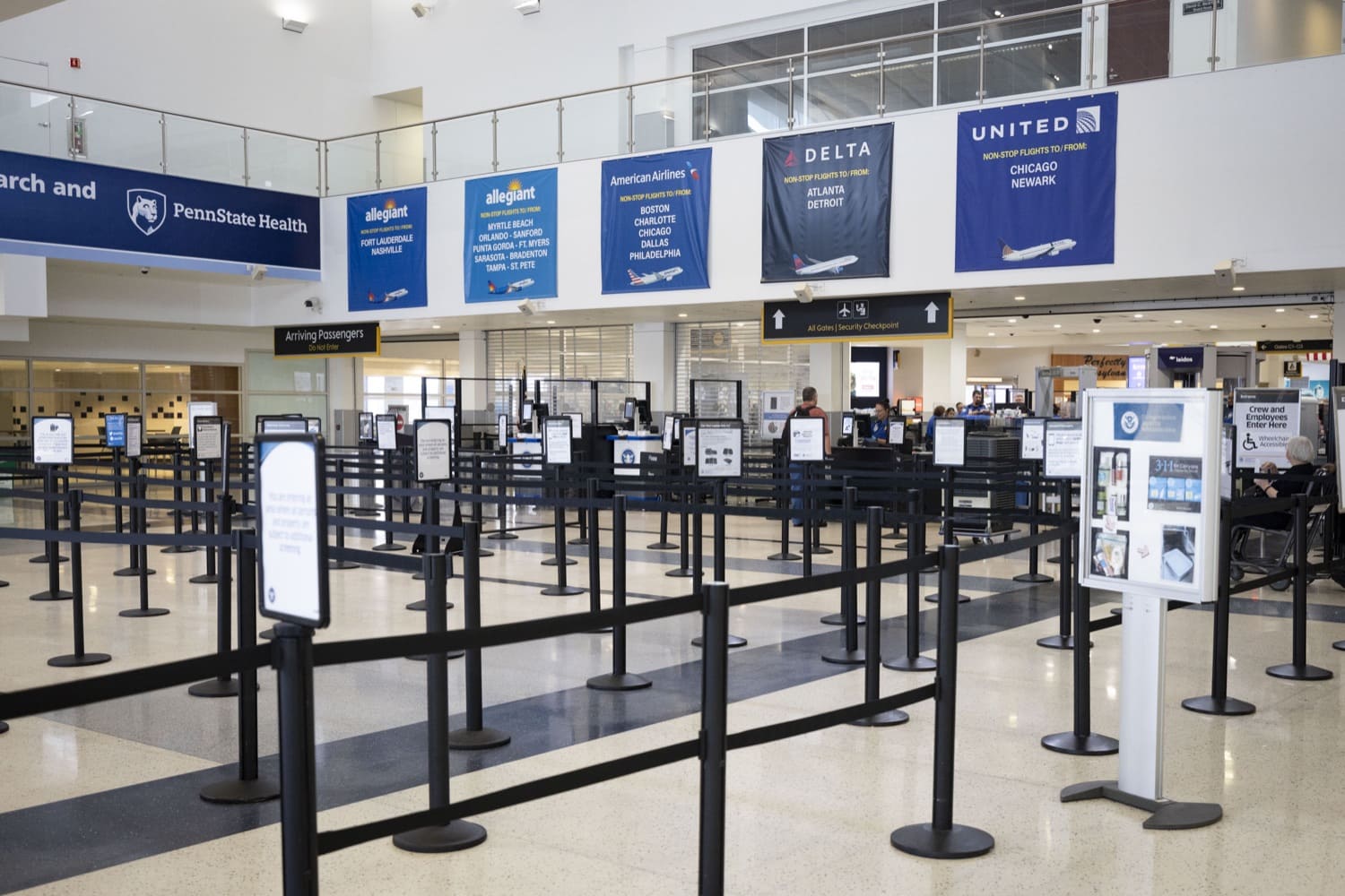 Several black stanchions line a security checkpoint inside a commercial airport. In the background, a TSA agent assists a customer.