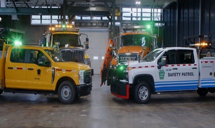 Two yellow PennDOT plow trucks, a yellow PennDOT crew cab and a white Pennsylvania Turnpike Commission crew cab sit next to each other in a maintenance garage with the green and amber lights flashing on the vehicles. 