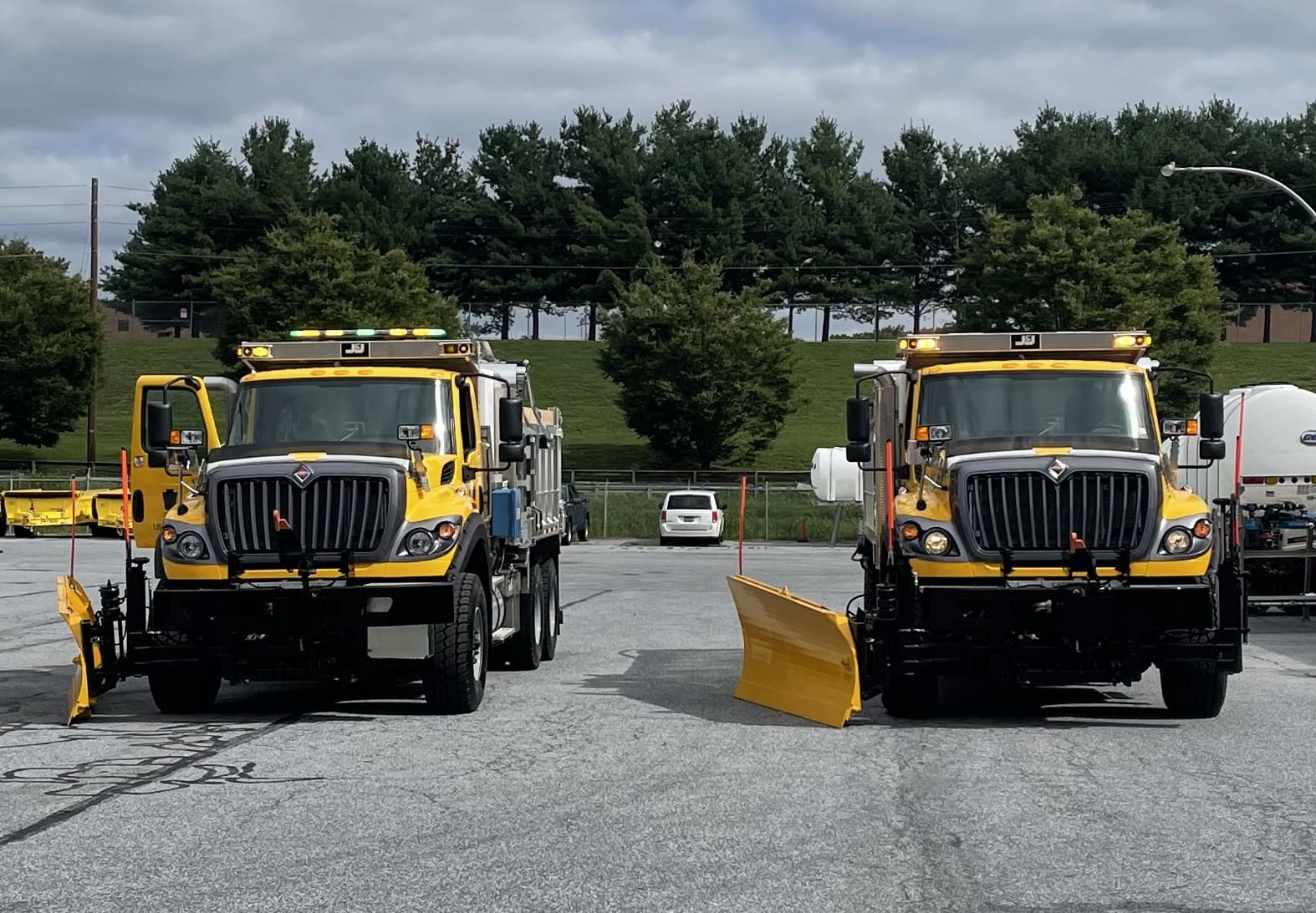 Two yellow PennDOT plow trucks sit in a parking lot with the green and amber lights flashing on the vehicles. 