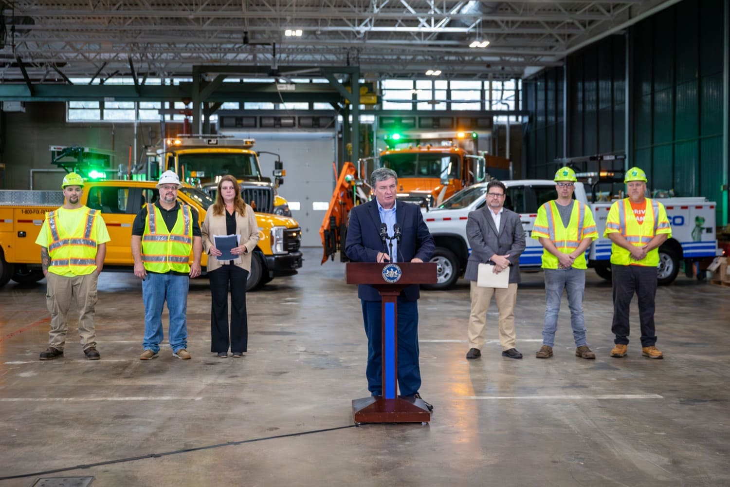 A man stands at a podium in the foreground. Six individuals stand behind him on either side of the podium. Four of the individuals are wearing yellow safety vests and hard hats and the other two individuals are wearing business attire. Behind them are four trucks affixed with green and amber lights on top. 