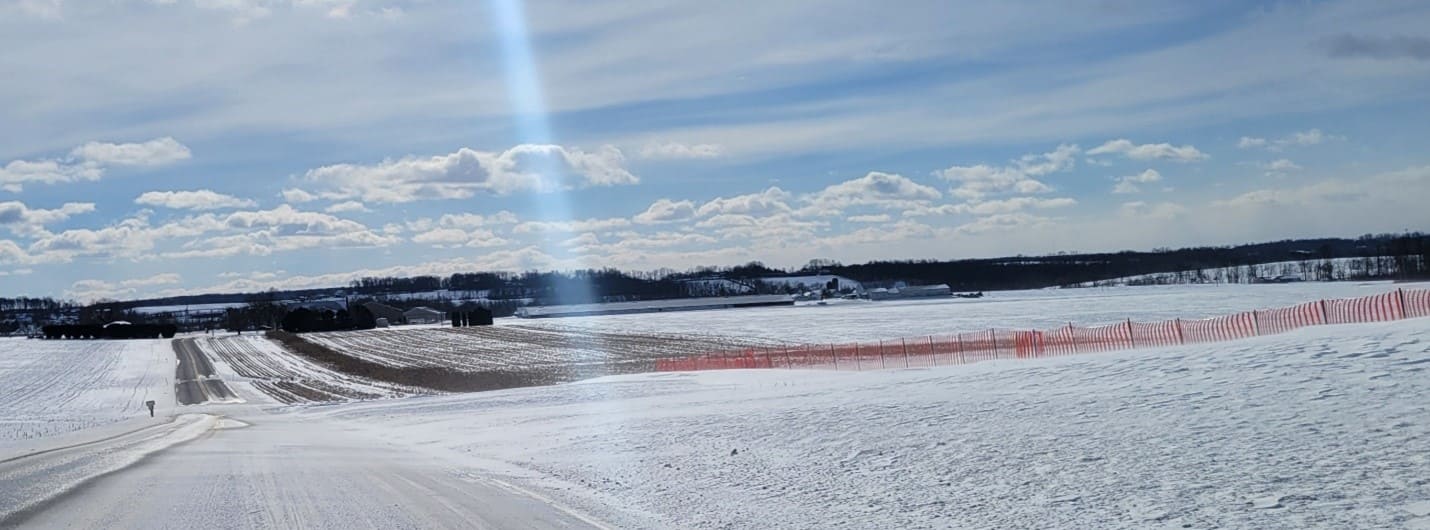 A photo of a snow-covered roadway with an orange snow fence and standing cornstalks on the right-hand side and an open field on the left-hand side. 