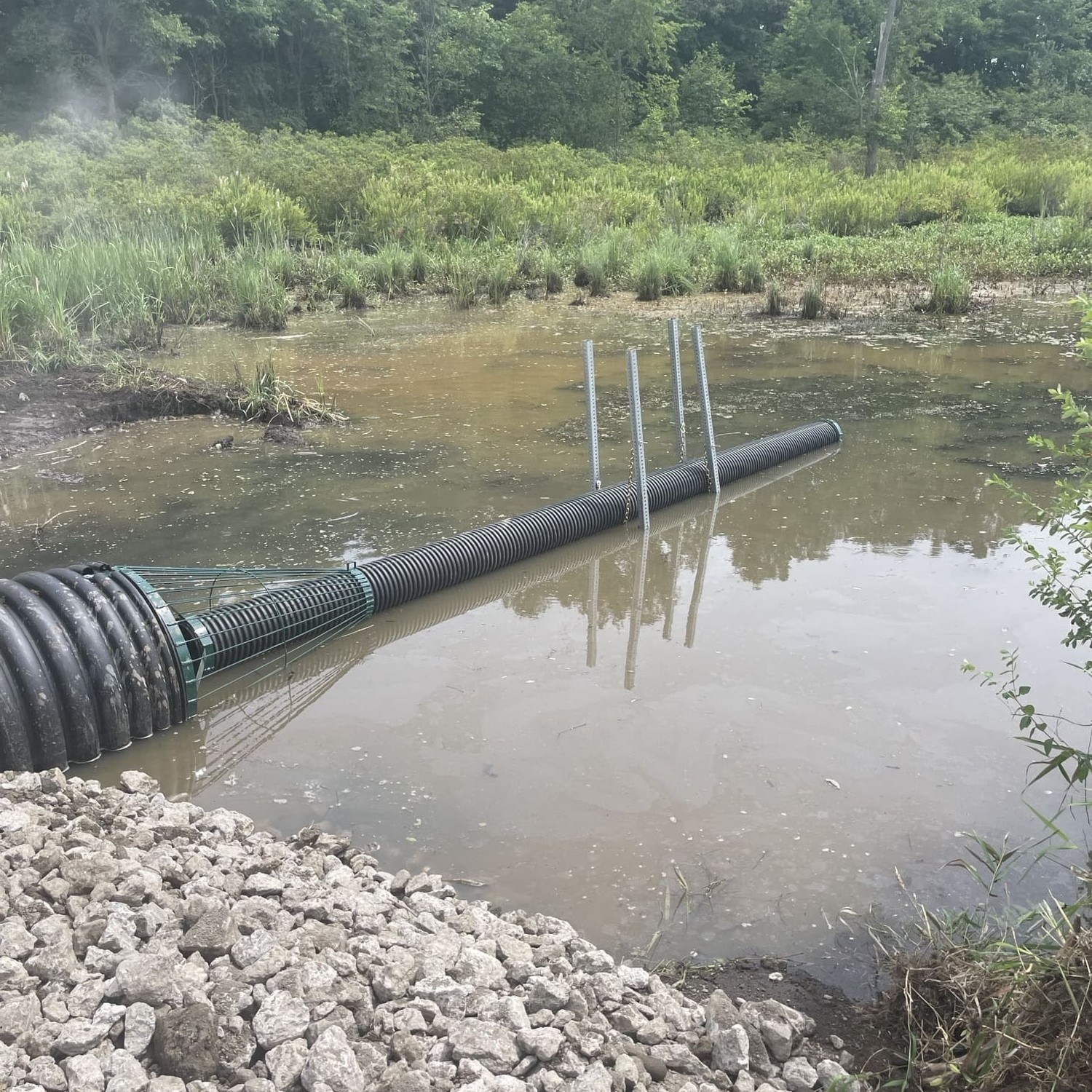 A cylindrical, metal cage with corrugated pipe running through the top of the cage sits atop water with rocks in the foreground and grass and trees in the background.