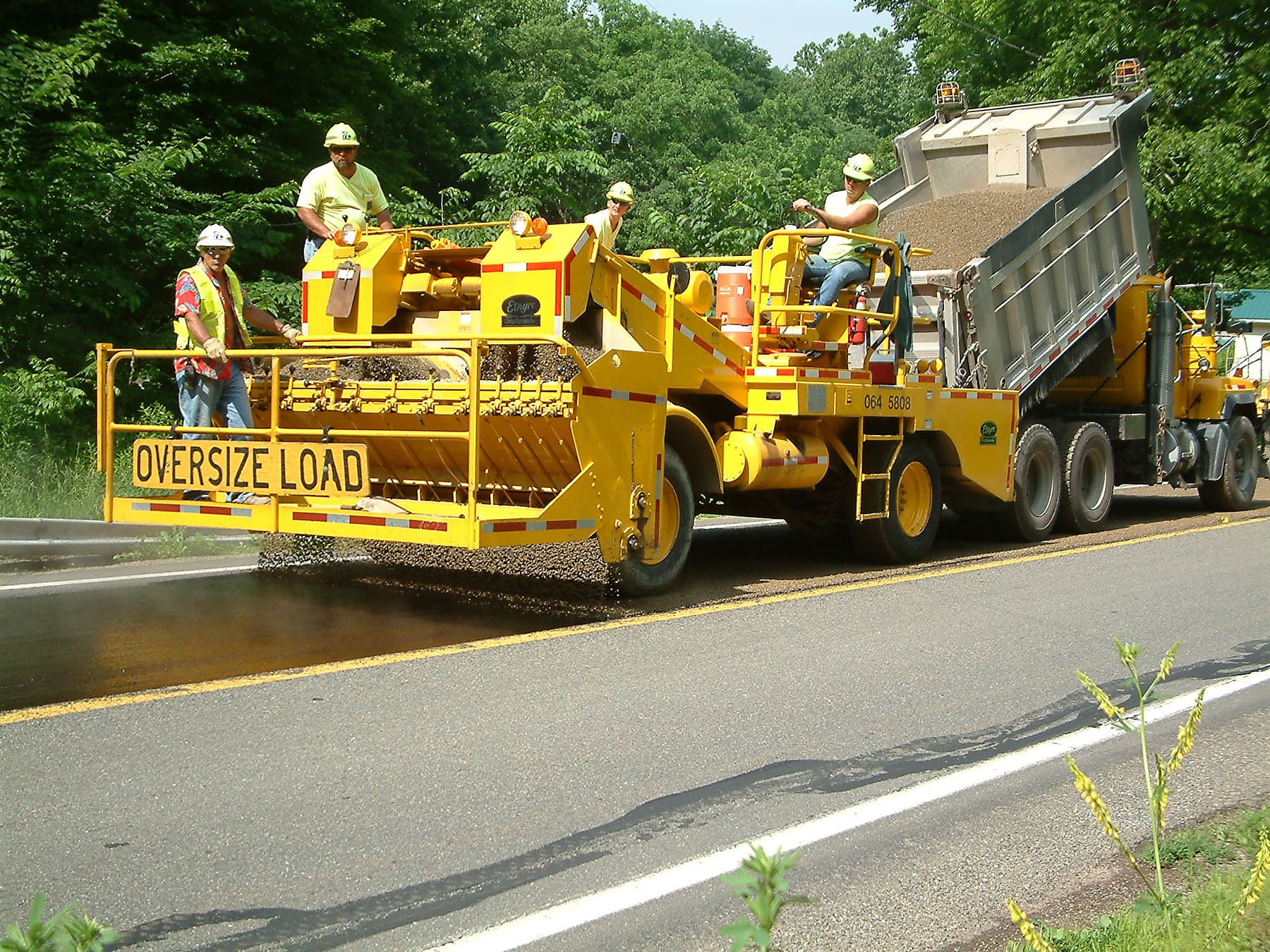Four crew members in yellow safety vests and white hard hats sit atop a yellow piece of equipment that is placing aggregate on the roadway. On the back of the piece of equipment are the words 'Oversize Load.' The piece of equipment is being pulled by a yellow dump truck.