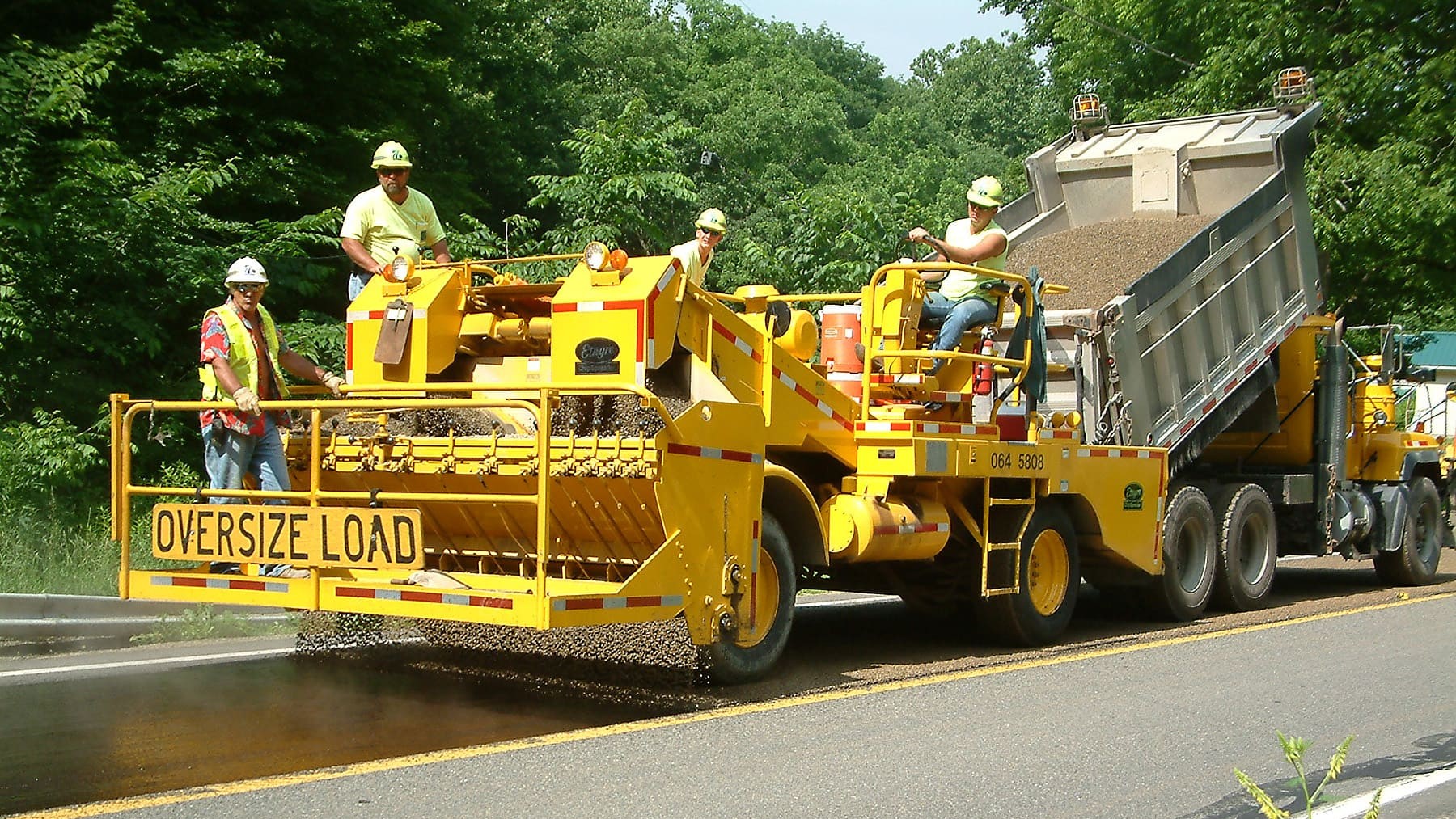 Four crew members in yellow safety vests and white hard hats sit atop a yellow piece of equipment that is placing aggregate on the roadway. On the back of the piece of equipment are the words 'Oversize Load.' The piece of equipment is being pulled by a yellow dump truck.