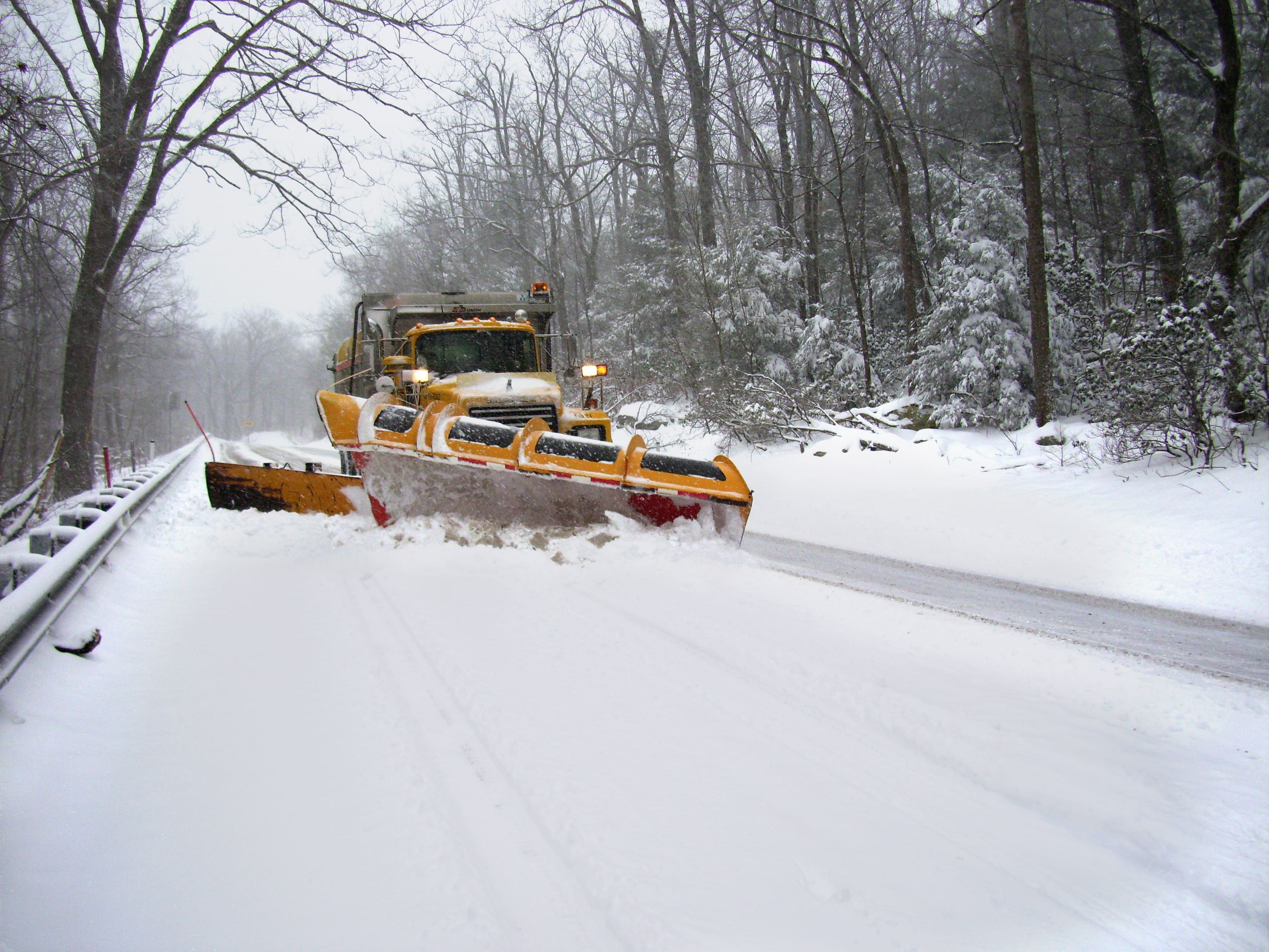A yellow plow truck plows snow on a rural roadway. Trees line either side of the road.