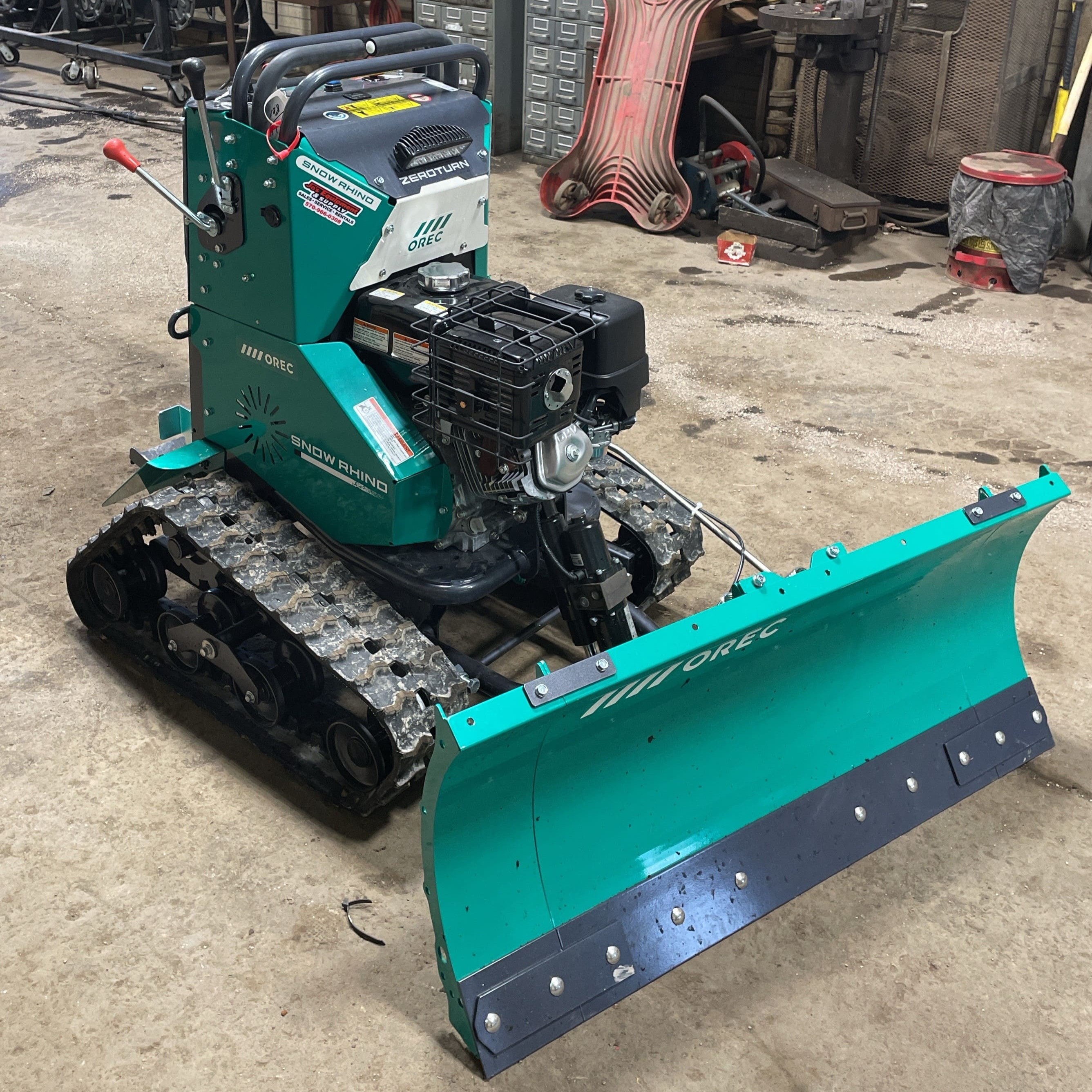 A small, green snowplow sits on a concrete floor in a maintenance garage. 
