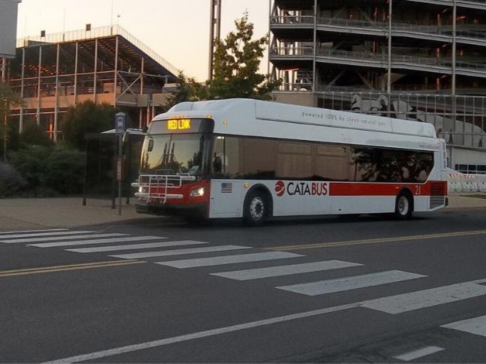 A white and red transit bus is parked at a transit stop outside a sports stadium. In the foreground is a crosswalk, and the stadium is in the background.