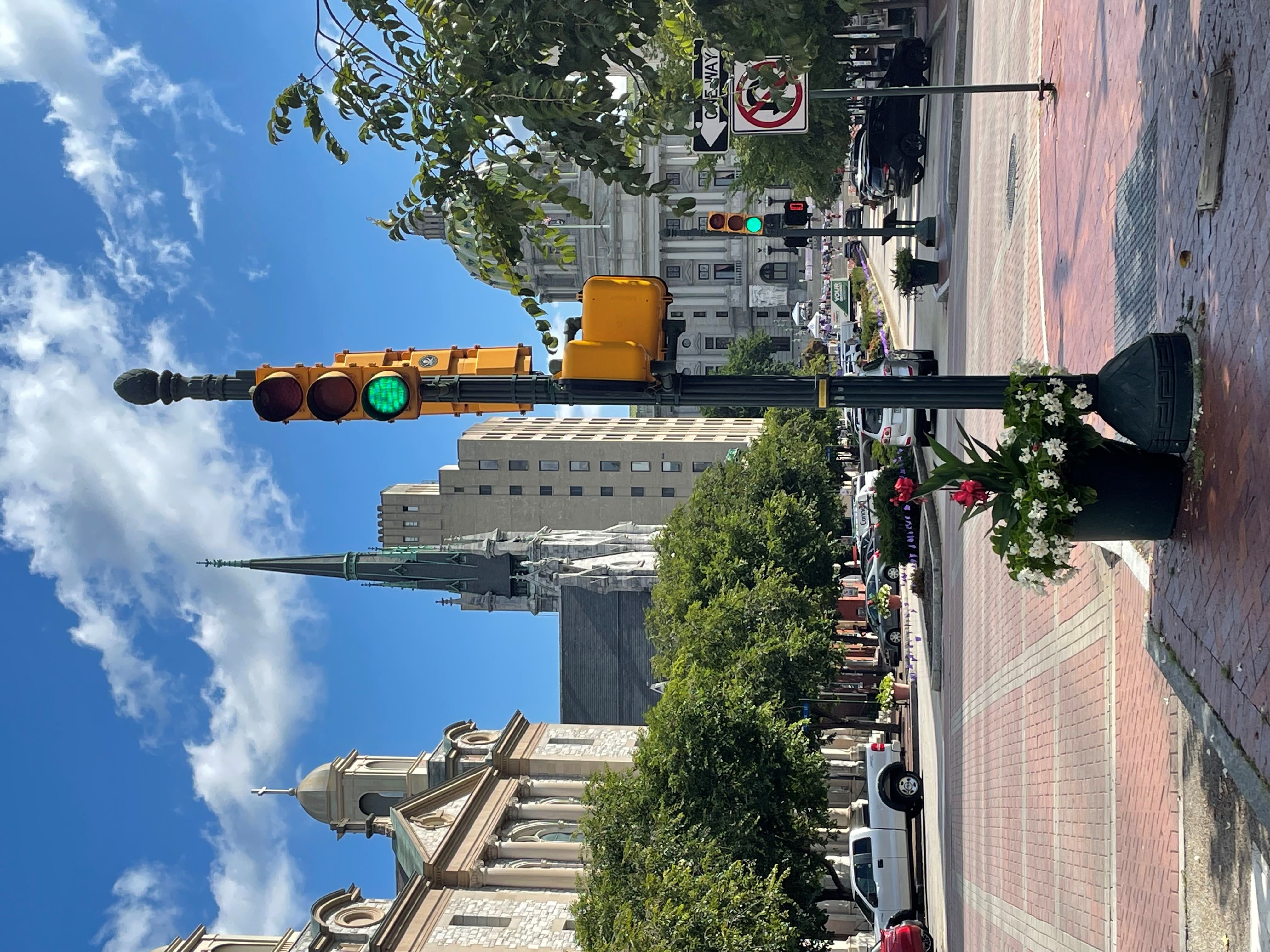 An image of a traffic signal on a street corner. The signal is showing green. In the background are several large buildings, including a church and the Pennsylvania State Capitol building. The sun is shining and there are blue skies with white puffy clouds. 