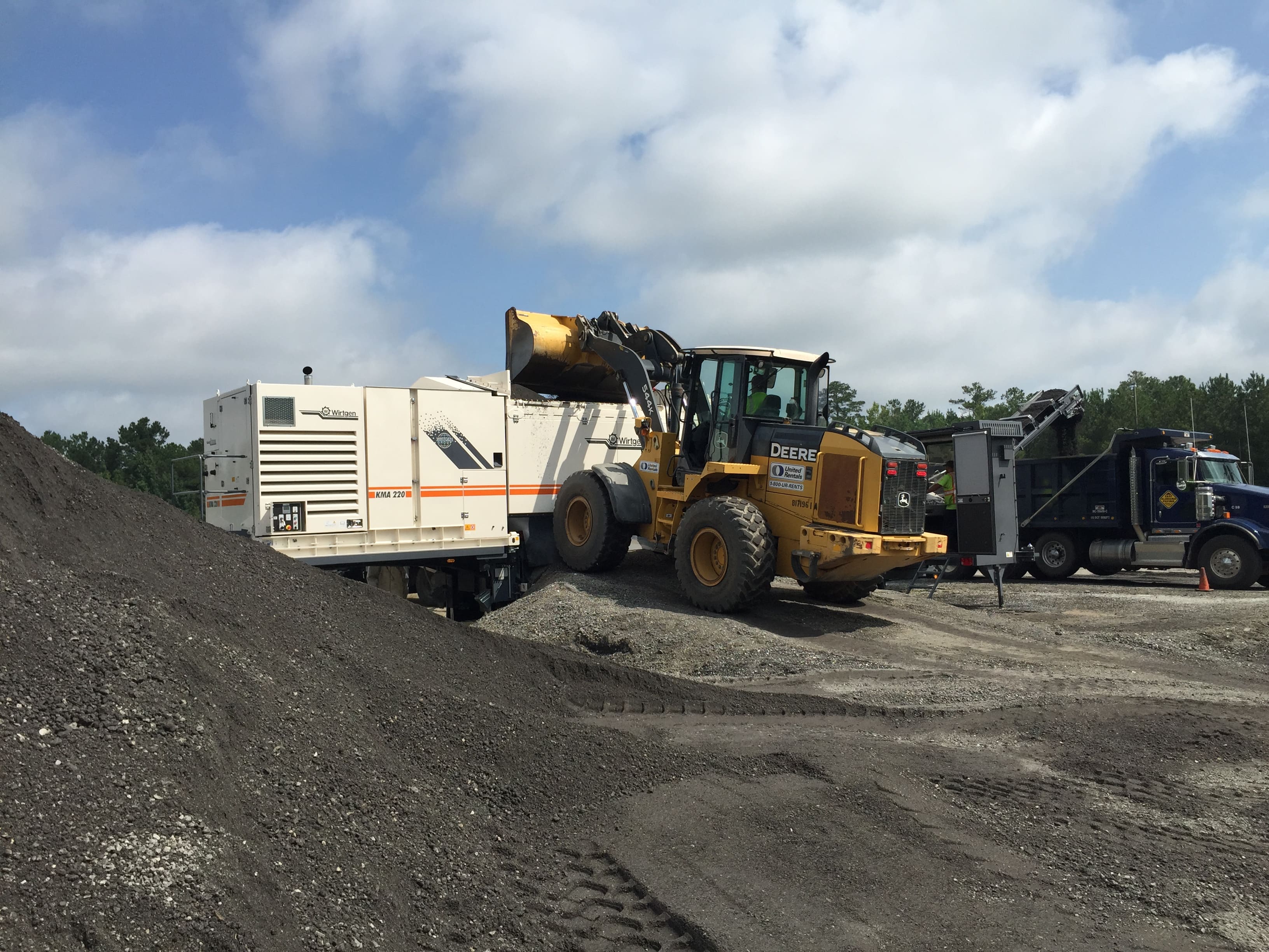An image of a worker operating a backhoe to place Reclaimed Asphalt Pavement (RAP) in a white, rectangle-shaped, metal mobile cold recycling plant where it is mixed with a liquid, such as asphalt emulsion or foamed asphalt, and a small amount of cement to create a new flexible asphalt base. 