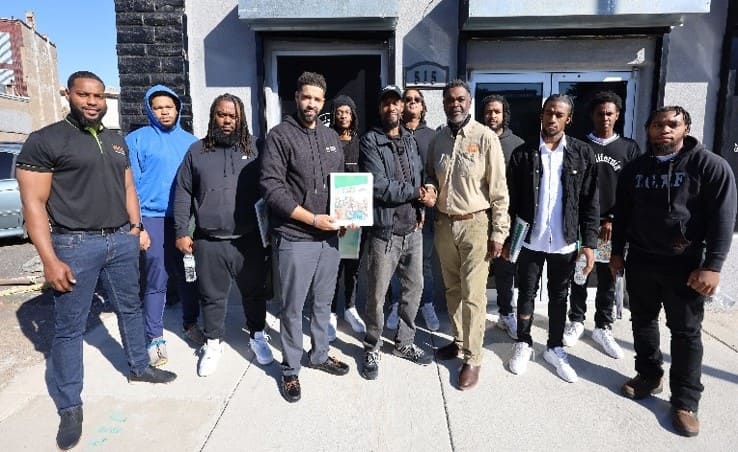 A group of 12 young men stand in front of a business. One man toward the middle holds an instruction manual, and two other men in the middle are shaking hands.