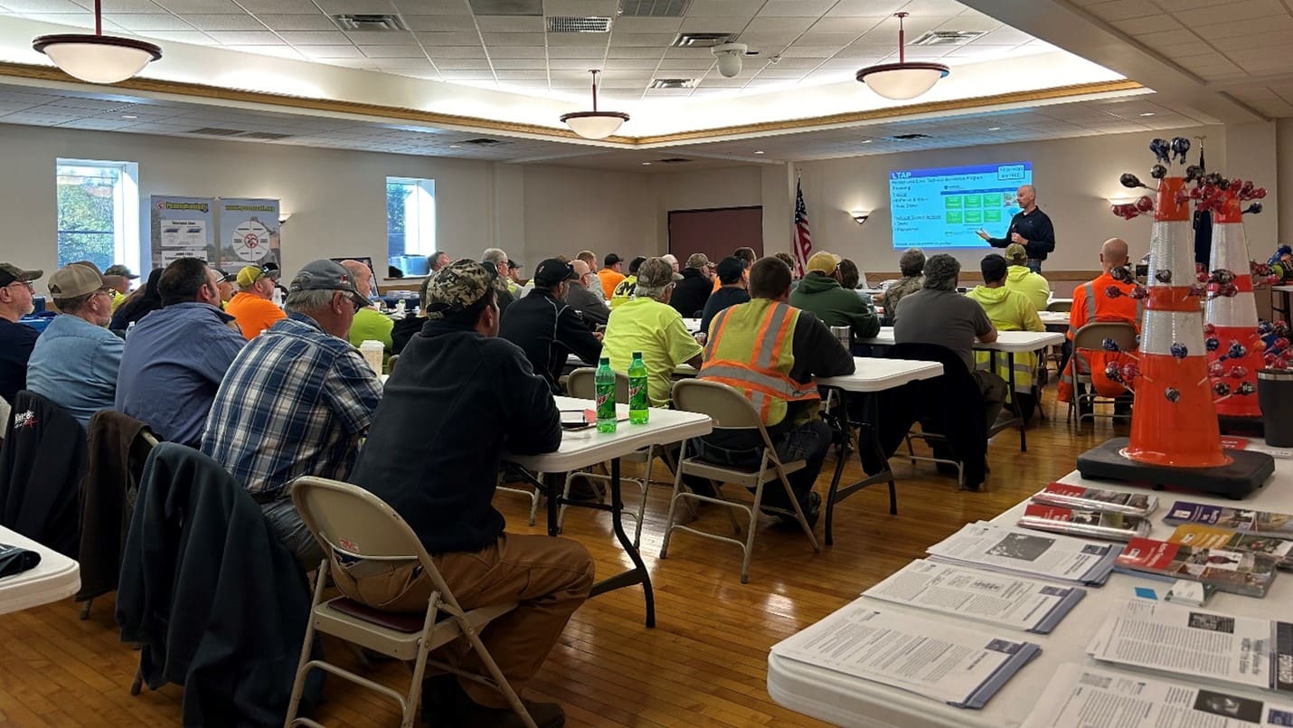 Several individuals sit at four rows of tables, while an individual stands in front providing training. An image is projected on the wall behind the instructor. Orange safety cones sit on a table in the foreground of the photo.
