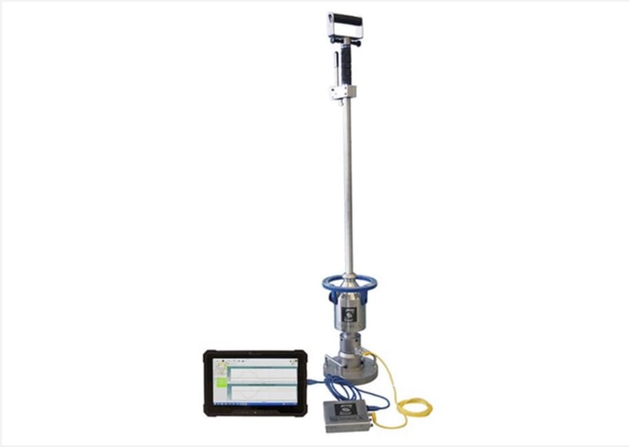 A metal device with a long handle and a calibrated weight on the bottom is connected to a tablet, which displays the measurements of surface deflection.