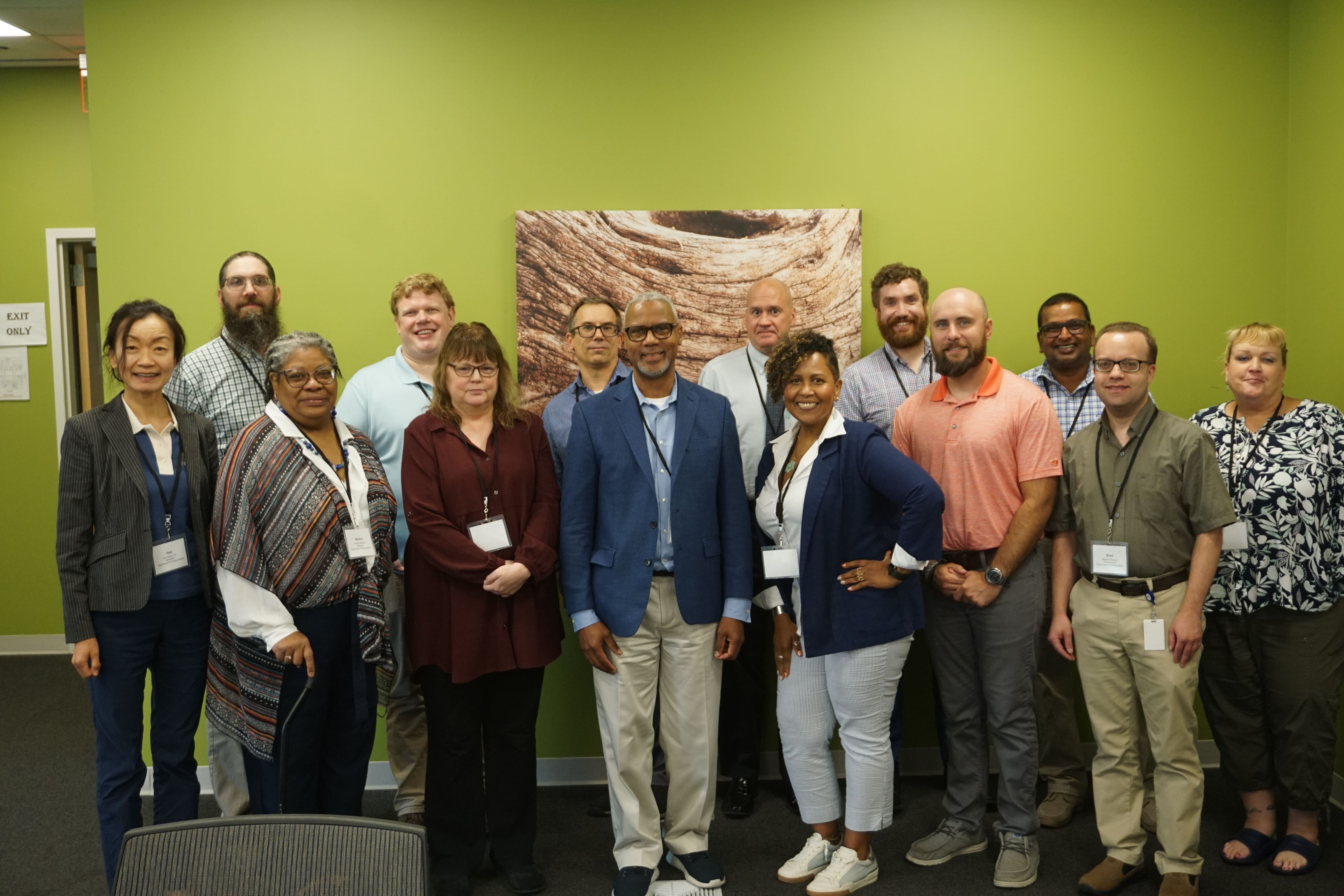 A group of 14 individuals stand together for a photo in front of a solid green wall with a photo of a rock formation hanging on the wall behind them. 