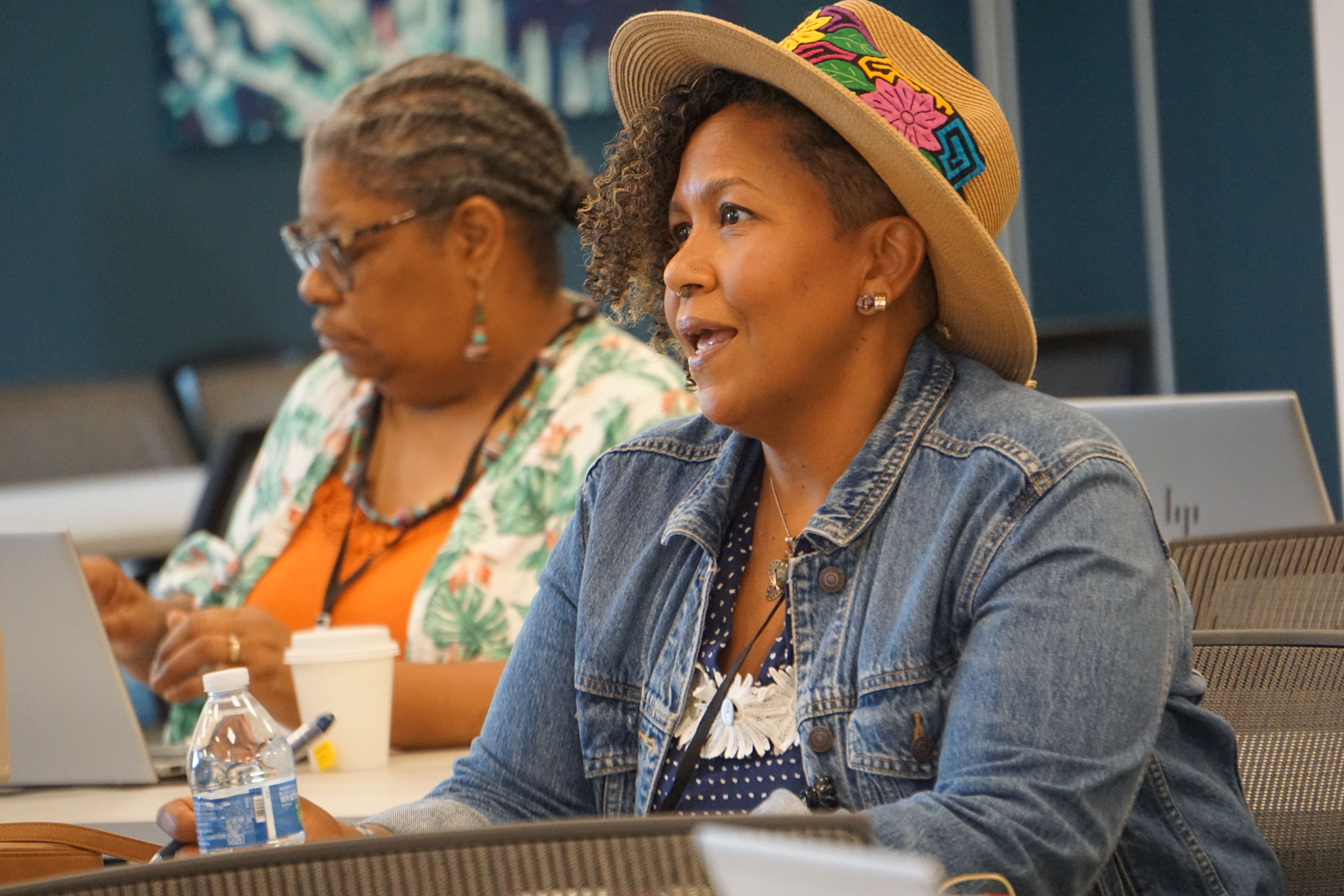 A woman wearing a blue jean jacket and hat sits at a table having a discussion, while another woman wearing an orange top and floral cardigan sits at the same table in the background.