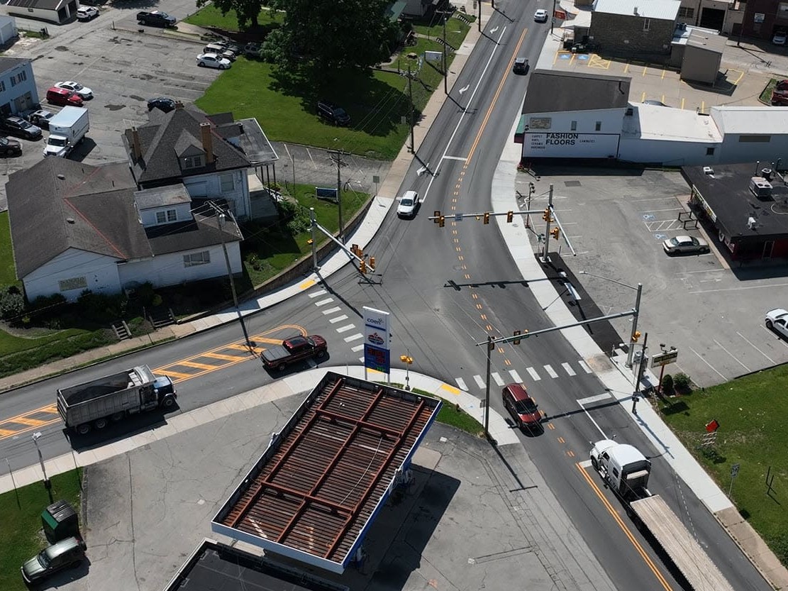 An ariel image of traffic navigating a T-intersection with traffic lights overhead and multiple houses and business surround the intersection.