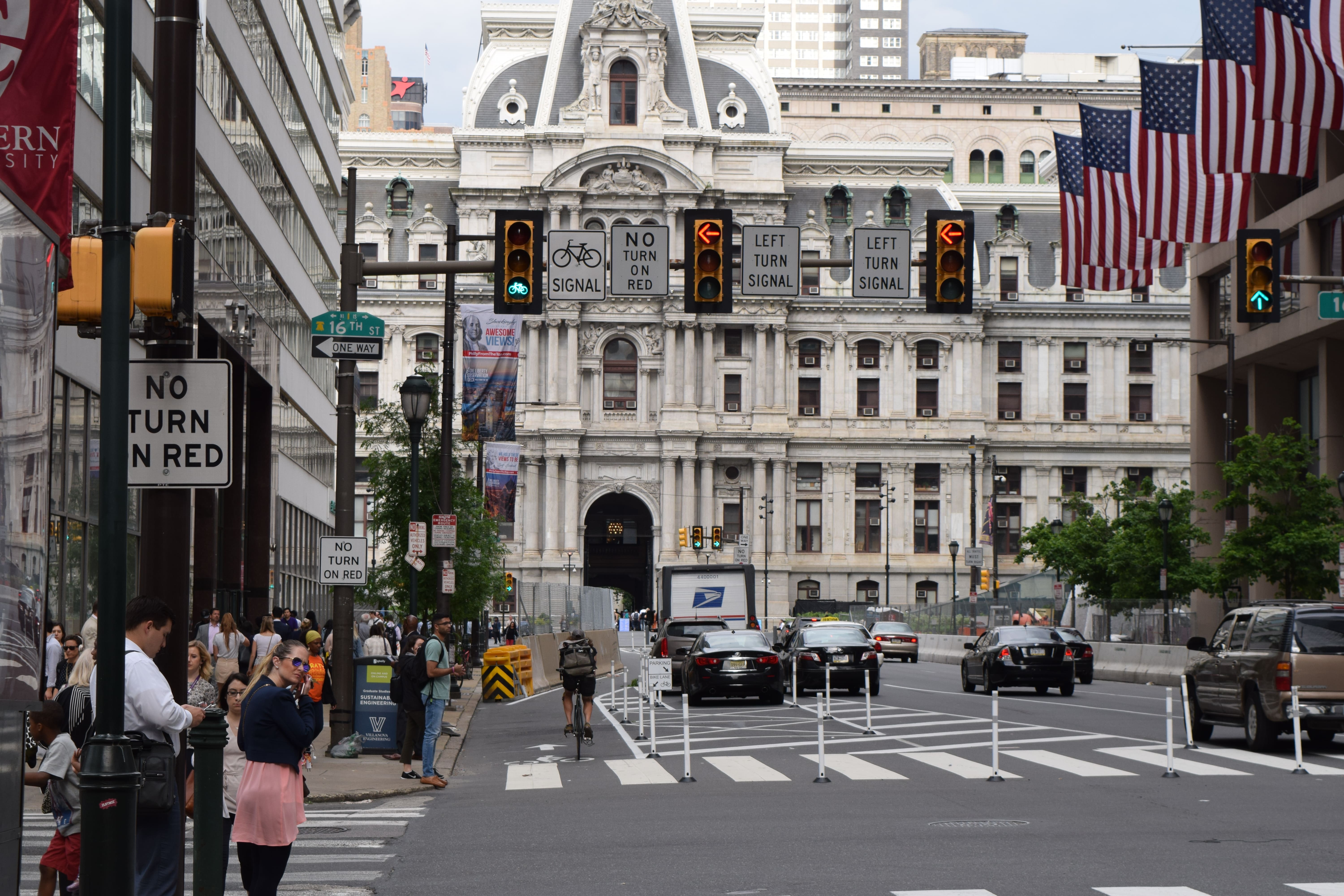 An image of a signalized intersection in Philadelphia in front of City Hall. Pedestrians stand on the sidewalk on the left. A bicyclist navigates a bike lane next to the sidewalk and several cars navigate multiple lanes of the city street.