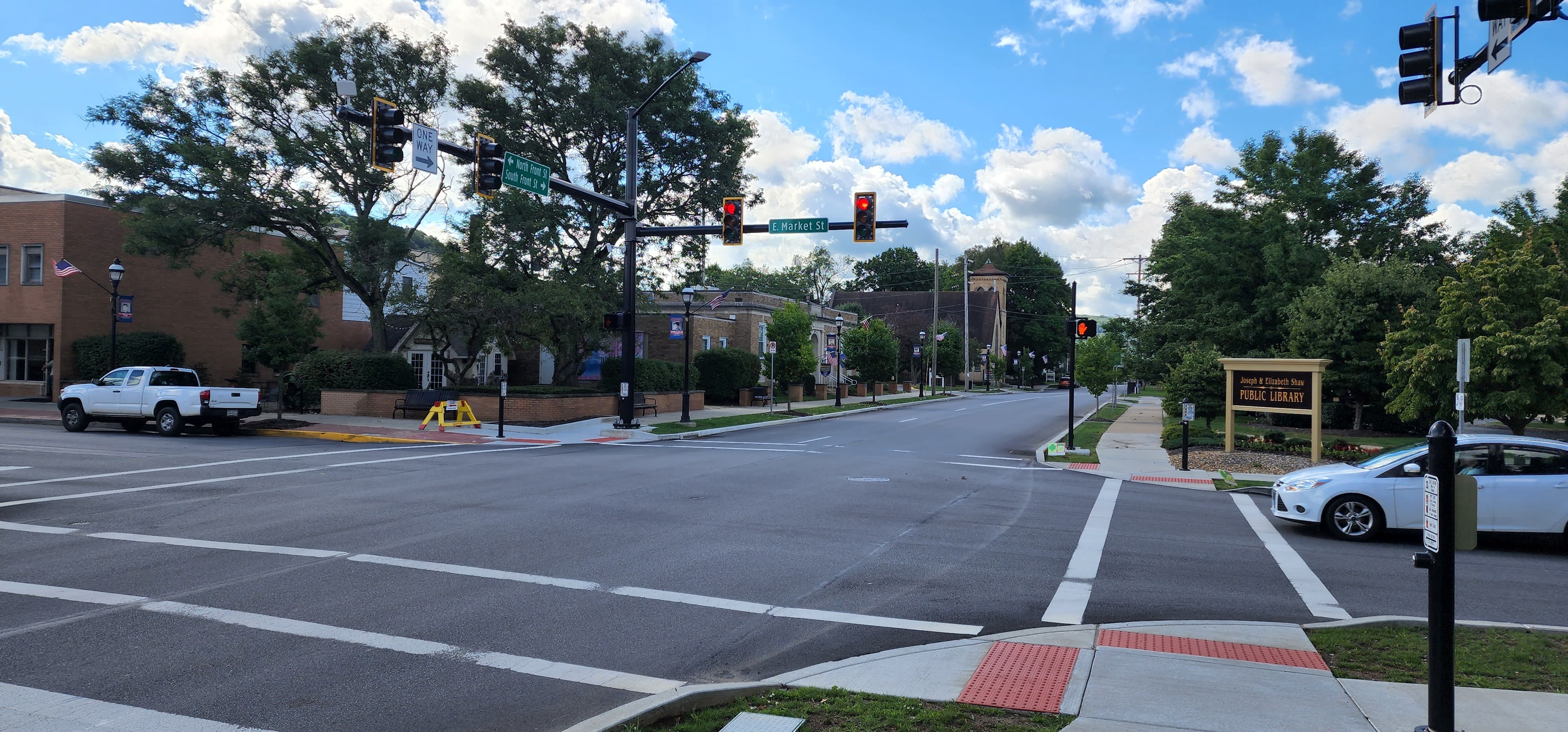 An image of a signalized intersection. A white car sits at the intersection on the right and a white truck sits parked on the other side of the intersection. Trees, blue skies and white puffy clouds are in the background.