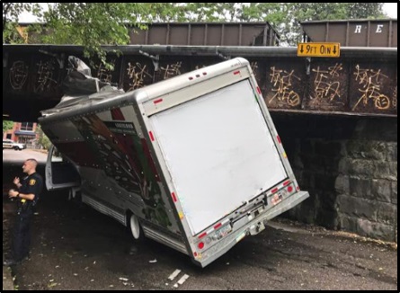 A rental truck is stuck under a railroad bridge with a height clearance sign showing nine feet, zero inches.