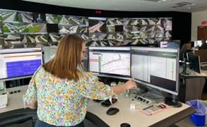 An individuals stands in front of several computer monitors, evaluating traffic conditions. A larger bank of traffic monitors hang on the wall in the background. 
