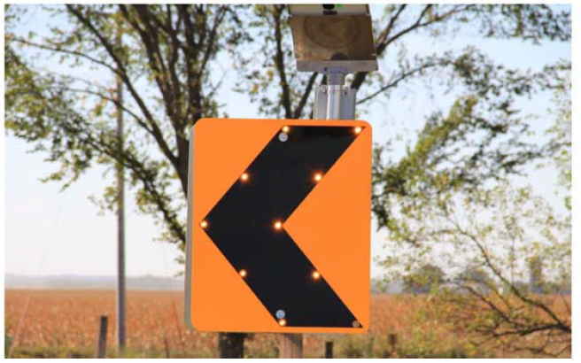 A yellow sign with a black arrow pointing to the left is affixed to a metal pole. Trees and a field appear in the background of the photo. 