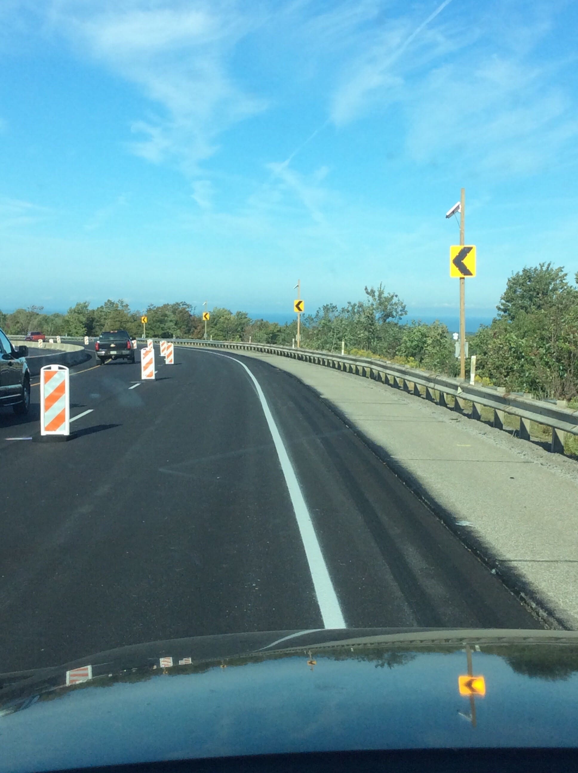An image of a car driving through a work zone with orange and white channeling devices on the left and a series of yellow signs with black arrows pointing left appear on the right on the other side of guardrail.