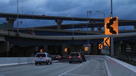An image of a cars driving on an interstate highway yellow flashing signs with black arrows pointing left appear on the right on the other side of concrete barriers. 