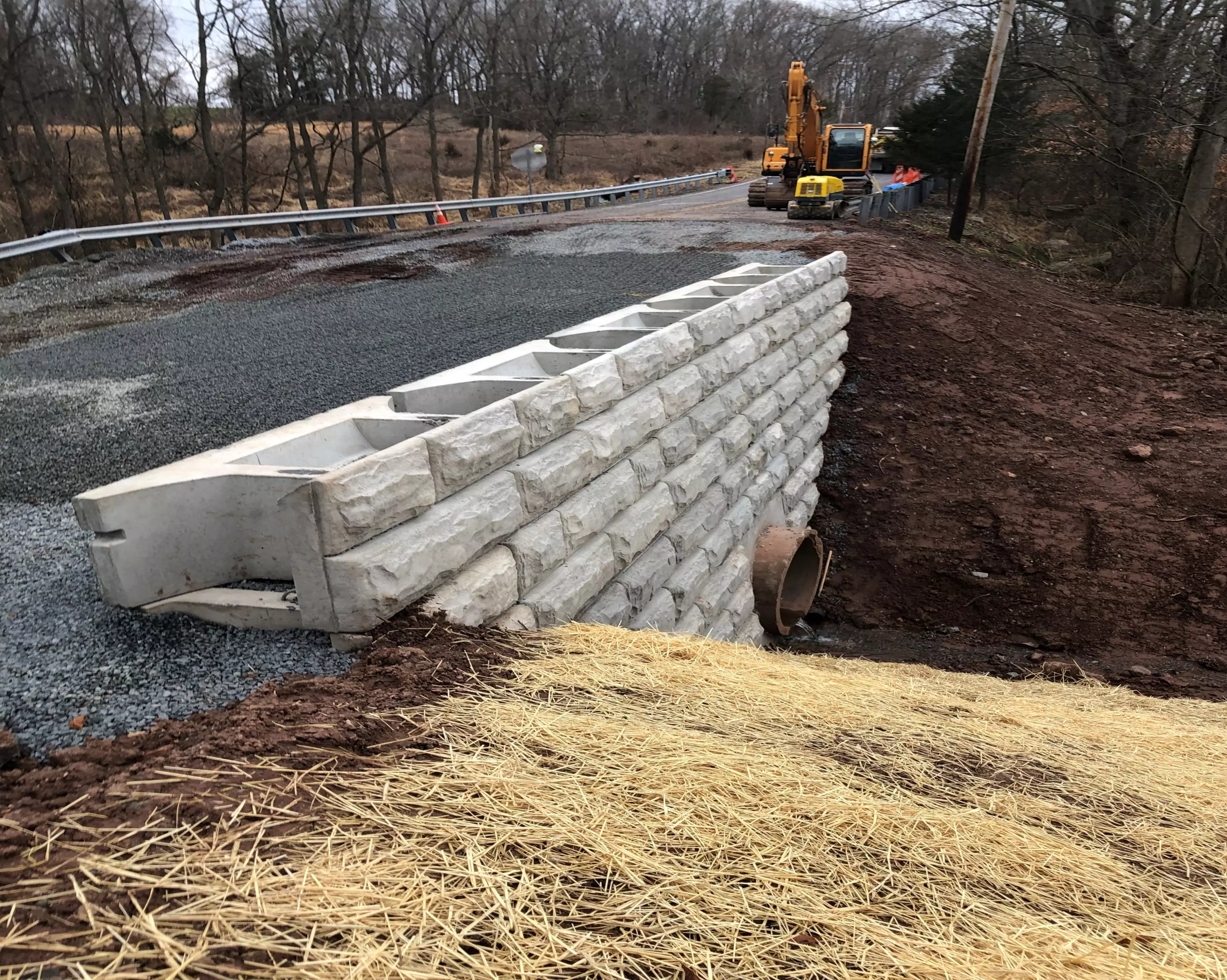 An image showing new retaining wall built on top of and surrounding a brown pipe outlet, with PennDOT construction vehicles in the background.