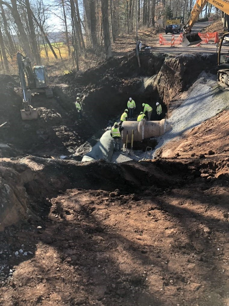 An image of PennDOT workers in yellow safety apparel and white hard hats working around the large pipe below the roadway.