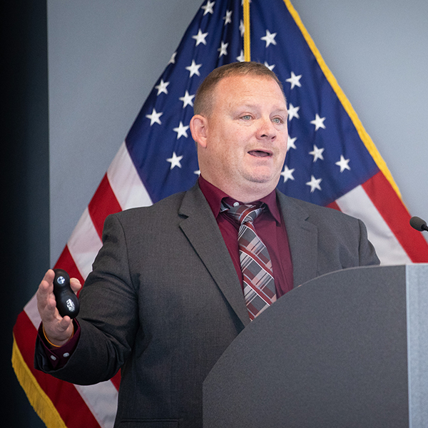 Harold Hill speaking at a podium, American flag in background.