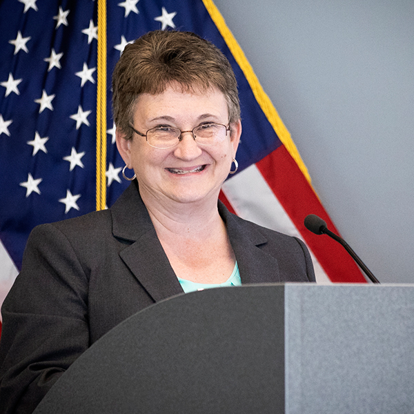 Karen Michael speaking at a podium, American flag in background.
