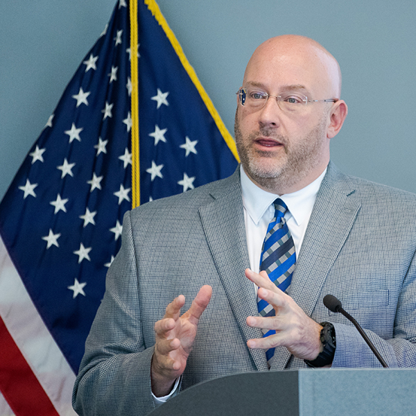 Rich Roman speaking at a podium, American flag in background.