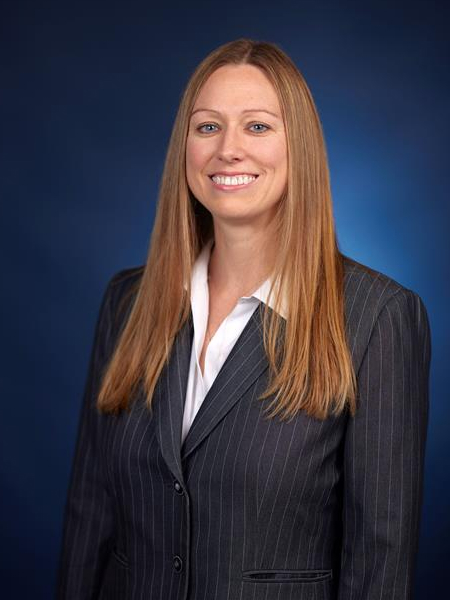 Professional portrait of Jennifer Sheffield wearing a dark pinstripe suit and white collared shirt, smiling in front of a blue background.
